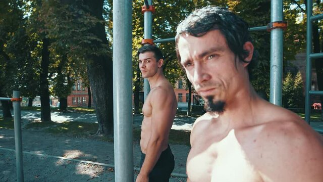 Portrait view of strong guy training outdoors. Serious caucasian unshaven man looking to camera while training with friend. Man posing with horizontal bars at background. Sport and training together