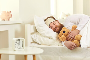 Happy grown-up man sleeping in good comfortable bed with cute teddy bear. Funny guy lying on white...