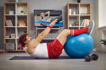 Funny chubby guy doing physical exercise with fitness ball lying on floor at home. Side view of man watching TV video lesson and having sports workout after gaining weight and getting fat in lockdown