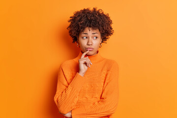 Fototapeta na wymiar Portrait of unhappy thoughtful dark skinned melancholy young woman feels upset looks aside with displeased expression thinks how to solve problem isolated over orange background. Monochrome shot.