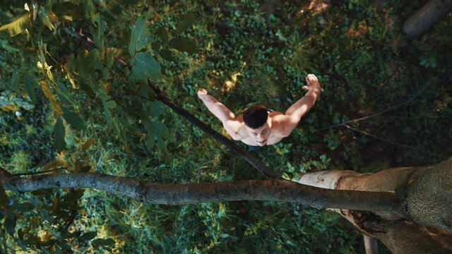 Male athlete is doing exercise with effort during workout on spring day. Top view of the young caucasian man is pulling up on the tree while training in garden. Guy practicing for health