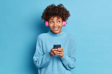 Fototapeta na wymiar People entertainment and leisure concept. Pleased African American woman bites lips wears stereo headphones listens audio track enjoys favorite melody dressed in sweater isolated over blue background.