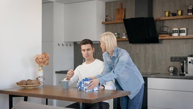 Young beautiful couple at home in the kitchen. A blonde woman gives her husband a gift for a holiday.