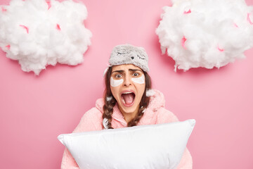 Emotional brunette young woman screams loudly keeps mouth opened wears blindfold nightwear holds soft pillow reacts on something amazing poses against pink background with fluffy clouds above