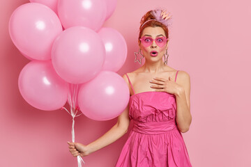 Shocked redhead woman stares surprisingly at camera reacts on aceepting congraulations from family and colleagues celebrates birthday holds bunch of inflated balloons isolated over pink wall