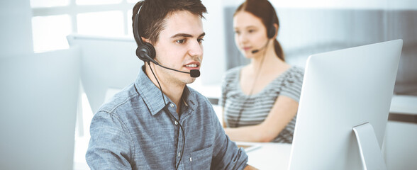 Casual dressed young man using headset and computer while talking with customers online. Group of...