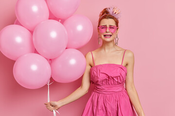 Obraz na płótnie Canvas Photo of frustrated redhead woman cries from despair poses in festive clothing with bunch of inflated balloons isolated over pink background. Bad holiday concept. This party sucks. Human emotions