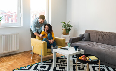 Happy couple using mobile phone together at home.
Loving couple relaxing together at home. Boyfriend showing smartphone to smiling girlfriend who is sitting on yellow armchair in living room