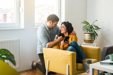 Cheerful loving couple looking in each other and laughing, woman holding mobile phone at living room. Embraced couple using smartphone together at home