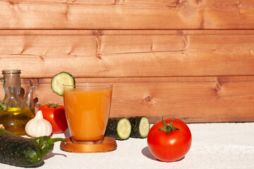 Concepto de un vaso de gazpacho en una mesa blanca con un fondo de madera y los ingredientes del mismo alrrededor del vaso para realizarlo.