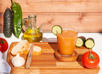 Concepto de un vaso de gazpacho andaluz con un fondo de madera en una mesa blanca con todos los ingredientes para hacerlo.