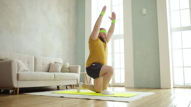 Fat man newcomer in sport is doing squats in lunges exercise standing on mat at home, side view. He is in yellow sportswear. Joke, mem, humor, parody. Workout, training, fitness, weight loss concept.