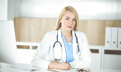 Woman-doctor at work while sitting at the desk in hospital or clinic. Blonde cheerful physician filling up medication history record form