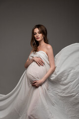 Pregnant girl in white flying cloth. Keeps belly. Grey background.