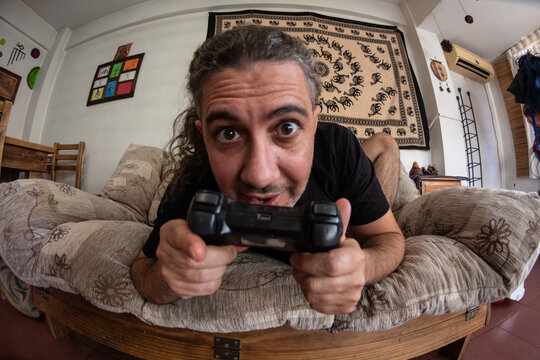 Hombre de pelo largo juega con su joystick mirando a c&aacute;mara sentado en el sill&oacute;n