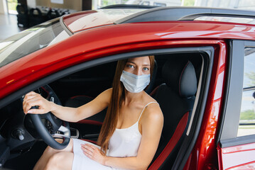 A young pretty girl inspects a new car at a car dealership in a mask during the pandemic. The sale and purchase of cars, in the period of pandemia.