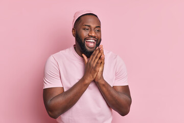 Positive dark skinned man keeps palms together has faith in something good being amused feels happy...