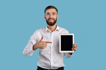 Young businessman pointing at tablet with empty screen on blue background, mockup for application or website design