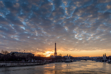 Naklejka premium Paris, France - February 12, 2021: CItyscape of Paris in winter. Ships and brigde over Seine river with Eiffel tower in background and dramatic cloudy sky