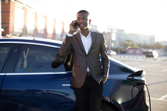 Lifestyle, transportation and ecology concept. Handsome young black man in business clothes, talking phone and waiting for his electric car recharging