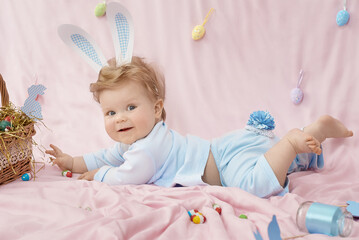 Cute little baby in bunny costume on pink blanket looking on us, he is wearing a blue bunny rabbit costume lying on his stomach near the basket of Easter eggs. Happy Easter time