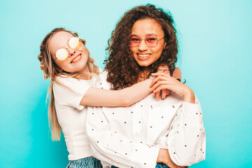 Two young beautiful smiling international hipster female in trendy summer clothes. Sexy carefree women posing near blue wall in studio. Positive models having fun in sunglasses. Concept of friendship