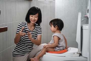 a mother comforts a pooping baby by watching a video on his cellphone on the toilet in the bathroom