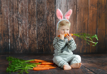 Cute young toddler boy wearing Easter rabbit ears chewing a carrot of a wooden background. Happy Easter time. Space for text