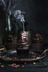 Several muffins or cupcakes with chocolate shaped cream at black table. Festive candle burns on a chocolate cake.