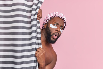 Shocked bearded dark skinned man hides behind shower curtain has amazed face expression applies...