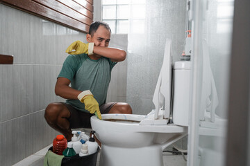 Sad man wearing gloves over his nose cleaning smelly toilets in the bathroom