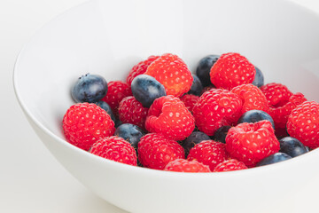 A close up detail of superfood mixed berries of raspberries and blueberries in a white bowl on a plain white surface and background.