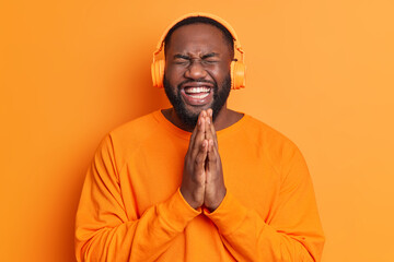 Overjoyed black bearded adult man keeps palms pressed together has upbeat mood laughs at something funny wears stereo headphones dressed in orange sweater isolated over bright studio background