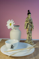 oriental still life with flower in a white vase, cup and a figure of a geisha on pink background