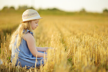 Obraz premium Adorable young girl having fun in a wheat field on a summer day. Child playing at hay bale field during harvest time. Kid enjoying warm sunset outdoors.