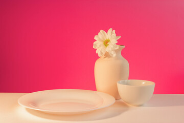 Minimalist still life with white vase, chrysanthemum flower, cup and plate on a vivid pink background