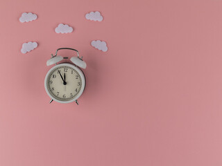 White alarm clock under the clouds on a pink background. Time: five minutes to twelve. Flat lay, top view. Copy spaсe. Concept: light mood despite the time.