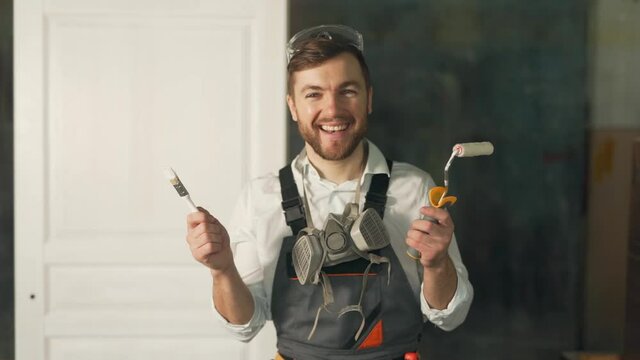 Cute man in working uniform of a painter holding a paint roller brush looking at the camera