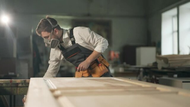 A handsome man in the form of a painter in a protective respirator paints a wooden product with a brush