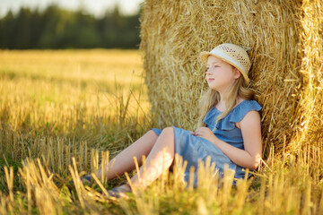 Adorable young girl having fun in a wheat field on a summer day. Child playing at hay bale field during harvest time. Kid enjoying warm sunset outdoors.