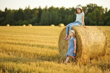 Naklejka na ściany i meble Adorable young sisters having fun in a wheat field on a summer day. Children playing at hay bale field during harvest time. Kids enjoying warm sunset outdoors.