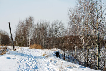 Snow-covered path in the village stretching into the distance. There is a cliff along the edge. A river flows at the bottom of the cliff. Winter landscape