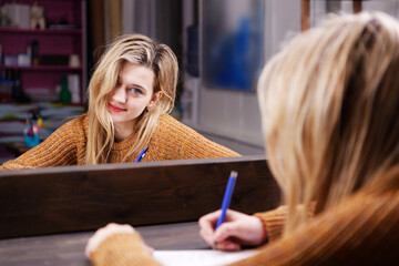 Young woman sits at a table in front of a mirror and writes something with a pen in a notebook....