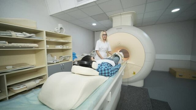 Woman doctor makes knee-joint MRI scanning. Young man patient on automatic table enters into a closed-type mri machine using noise isolation headphones. Modern equipment, coil on the patient's knee