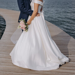 Naklejka na ściany i meble Bride and groom embrace on a wooden pier by the sea, the bride has a very long veil to the ground, the groom is dressed in a dark blue suit.