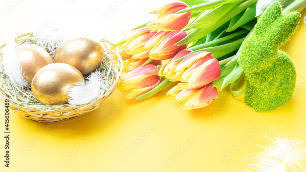Wall mural golden eggs in basket with spring tulips, white feathers on pastel yellow background in happy easter