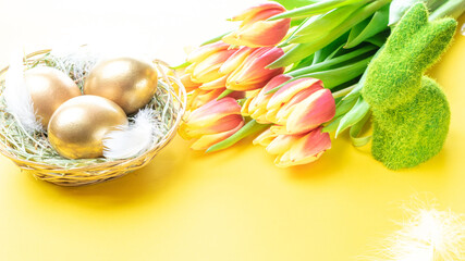 Golden eggs in basket with spring tulips, white feathers on pastel yellow background in Happy Easter decoration. Foil minimalist egg design, modern design.
