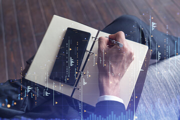 A trader in formal wear writing down some quotes to research stock market trends using smart phone for right investment solutions. Wealth management concept. Hologram Forex chart over close up shot.