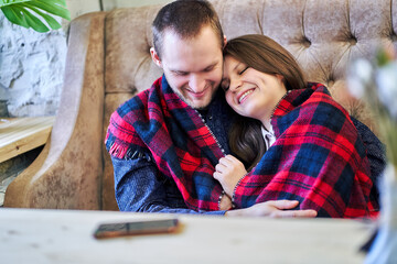 Lovers, wrapped in one blanket, hug while sitting on the couch.