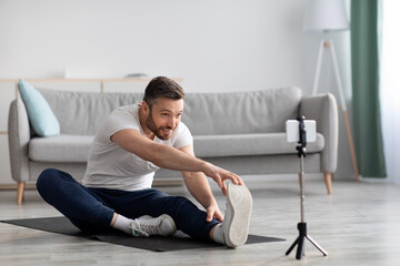 Cheerful sporty man stretching on fitness mat, broadcasting with smartphone
